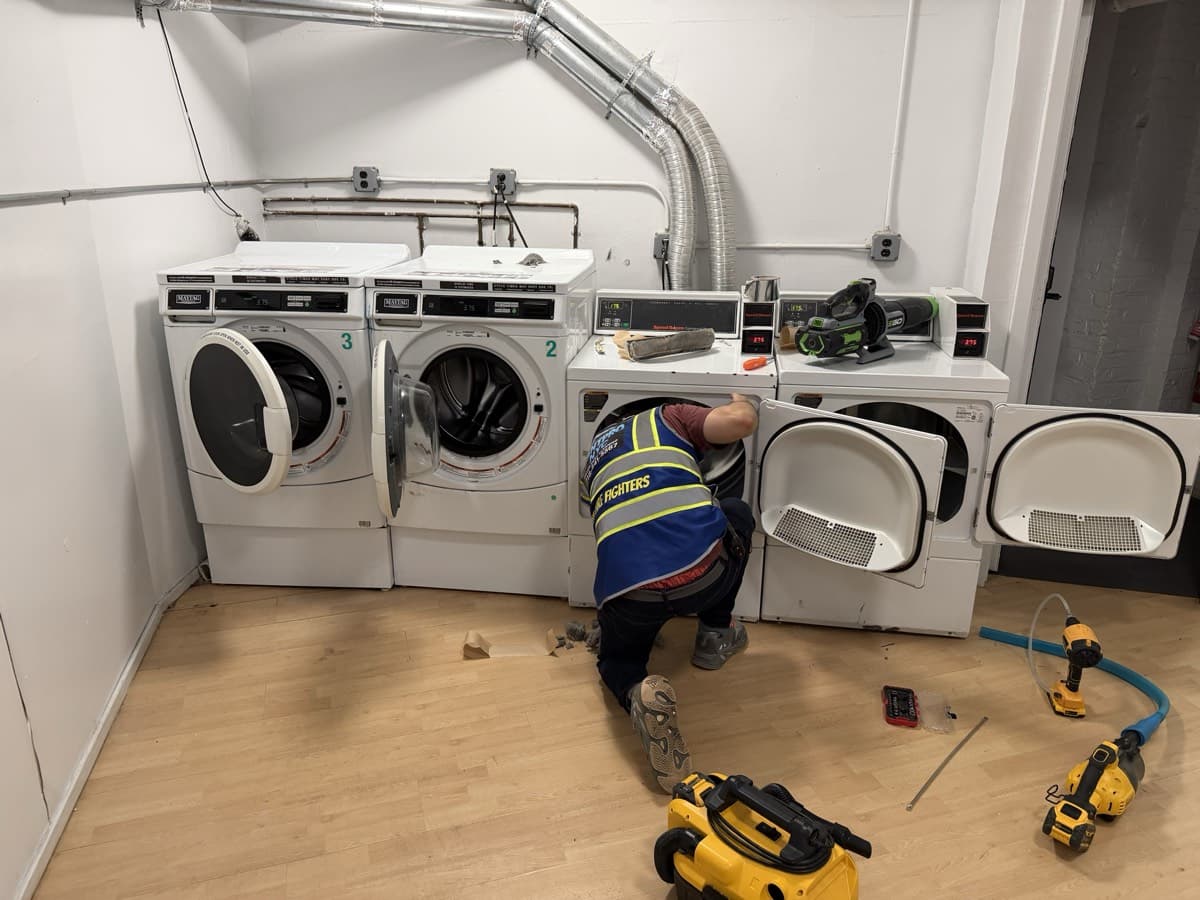 Vent Pro NYC technician cleaning a dryer vent in a building's shared laundry room lined with commercial washers and dryers